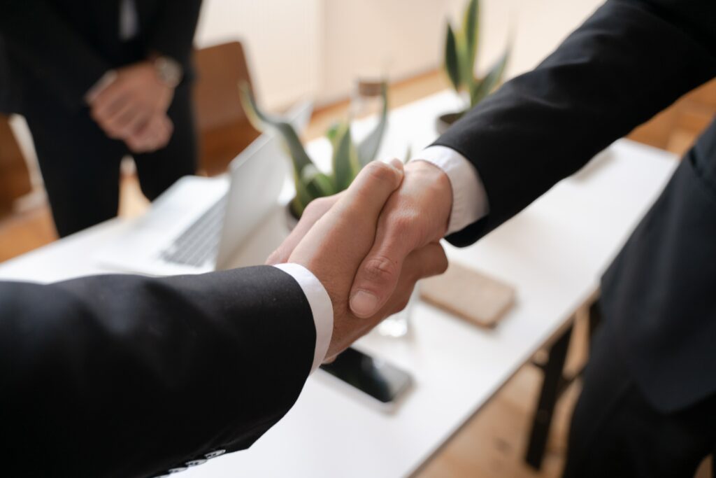 In a close-up view, men dressed in business suits engage in a handshake, while another man is seen in the background near an open laptop. The firm grip and professional attire signify a formal business interaction, while the presence of the laptop suggests a meeting or collaboration.
