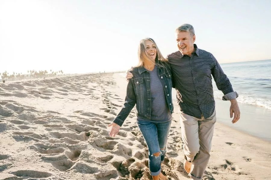middle-aged man and woman stroll barefoot along a sandy beach on a sunny day. The warm sunlight illuminates the scene, creating a tranquil and inviting atmosphere.