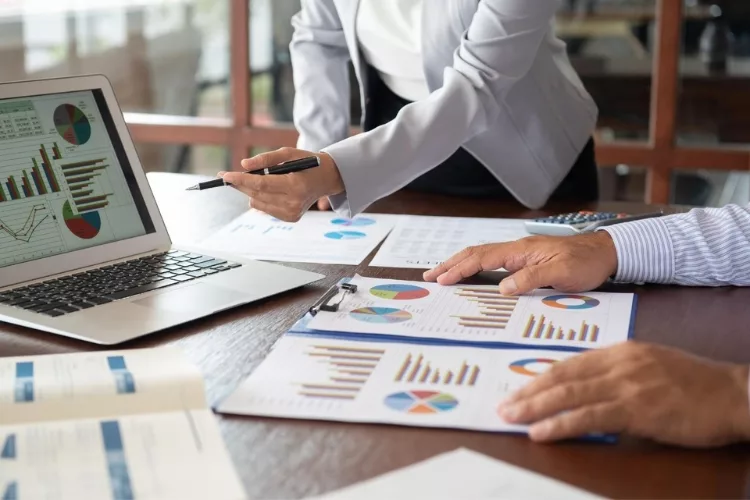A close-up image showcases the hands of a woman and a man as they carefully review financial documents. The laptop in front of them displays additional graphs, aiding in their analysis.
