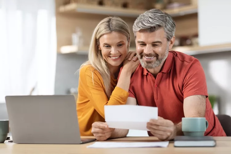 A middle-aged man and woman stand in their kitchen, focused on a piece of paper in their hands. With a laptop open on the table, they engage in thoughtful discussion and review.