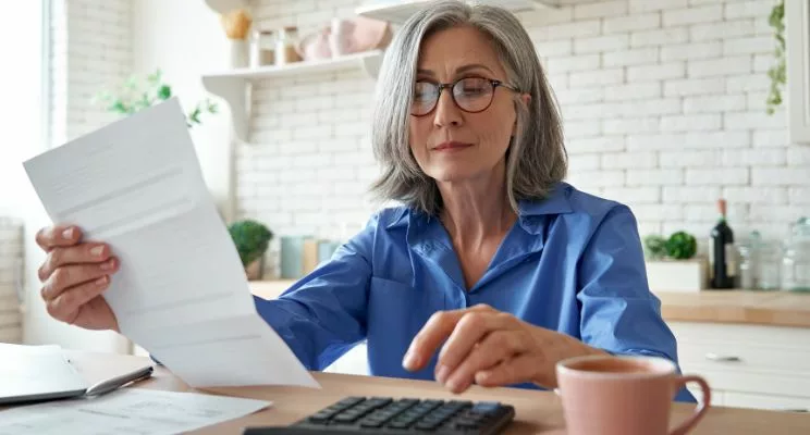 A woman wearing a blue shirt, seated at a kitchen table, focused on typing numbers into a calculator, depicting financial calculations or budget management.