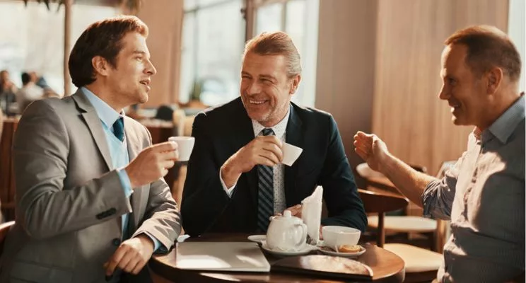 Three businessmen sitting at a round table, enjoying coffee, and engaged in conversation in a well-lit room, conveying a professional and collaborative atmosphere.